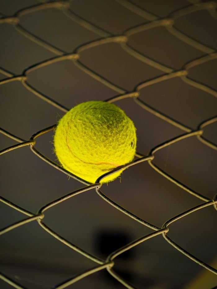 Menu A close-up of a yellow tennis ball wedged in a chain link fence.